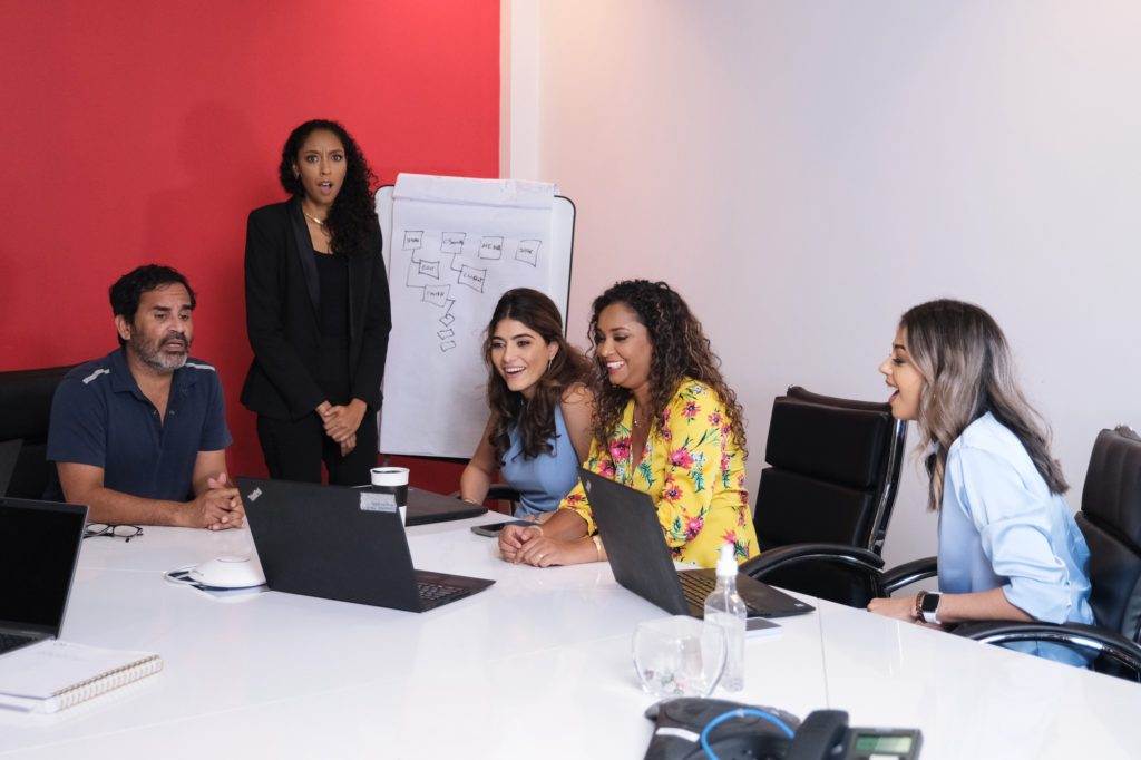 Five people in a meeting room are gathered around a table, looking at a laptop screen. A whiteboard with a flowchart is in the background.